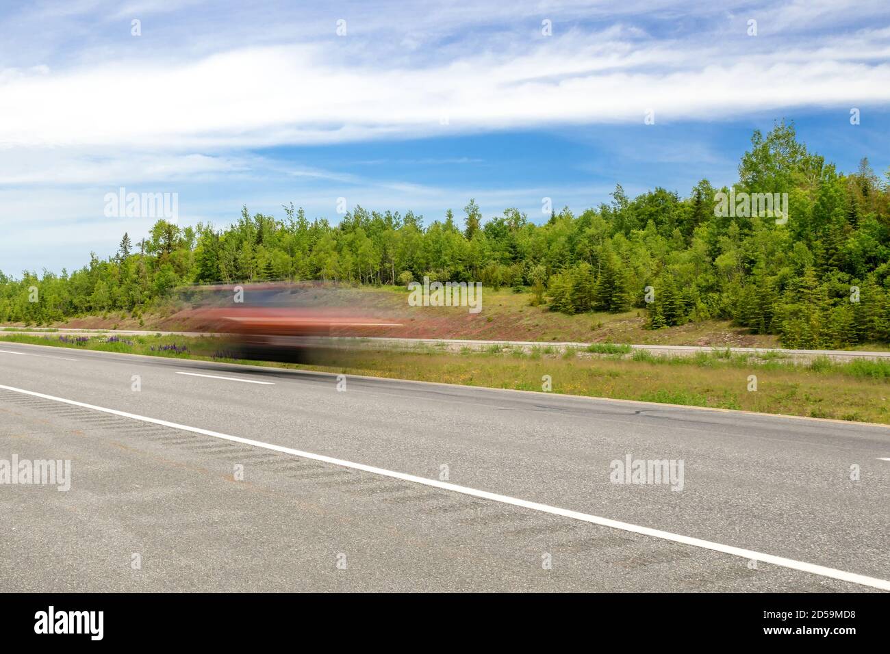 Fast moving red car on a highway. Car is just a blur due to long exposure. Trees in background, partially cloudy sky. Stock Photo