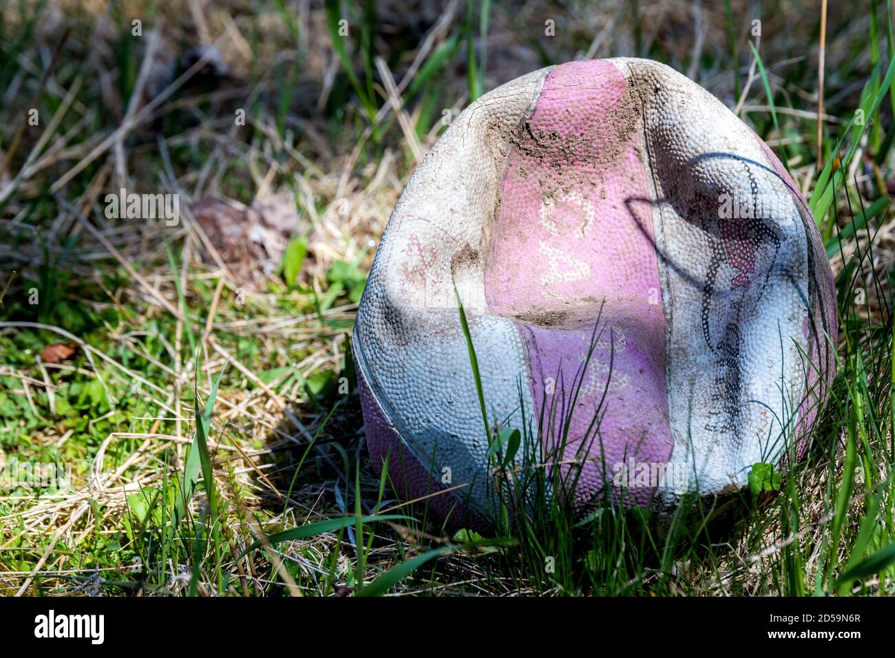 An old, faded, and deflated basketball lying in grass. Part shade and part sunlight on the ball. The ball is also dirty. Stock Photo