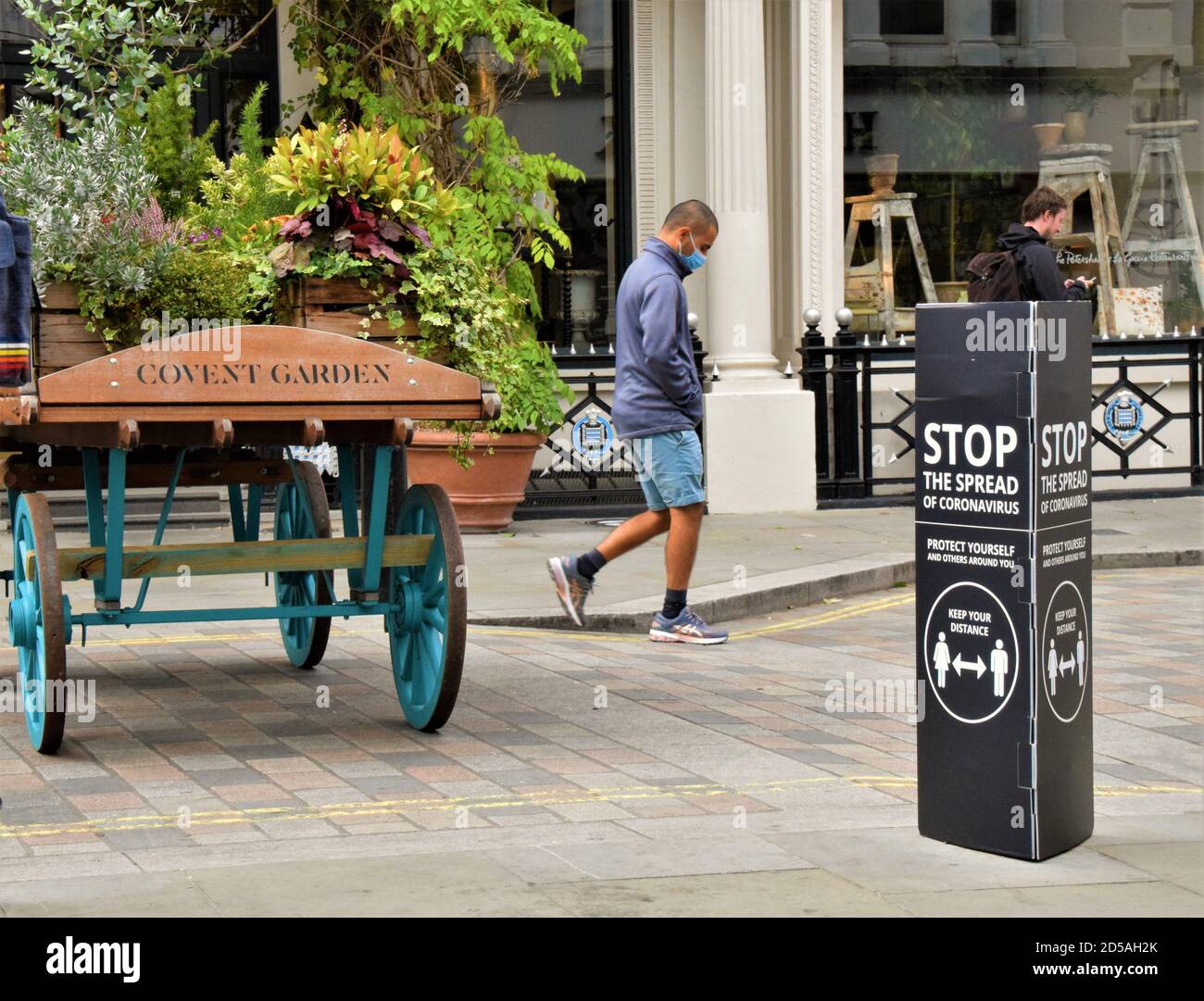 A man with a protective face mask walks past a Stop The Spread Of Coronavirus social distancing street sign in Covent Garden, London Stock Photo