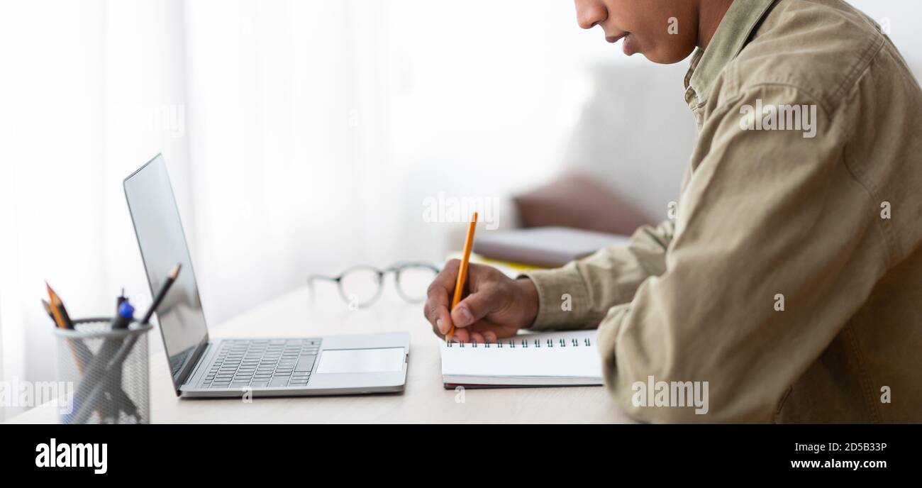 Web-based training. Closeup of black male student writing in copybook near laptop at home, banner design with copy space Stock Photo