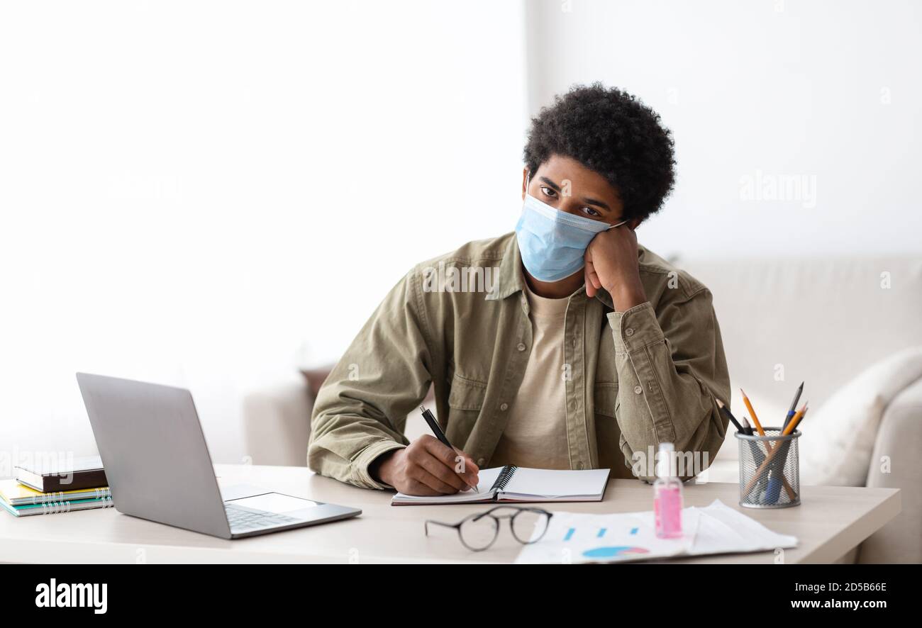 E-learning during coronavirus. Bored black teenager in safety mask writing in notebook near laptop computer at home Stock Photo