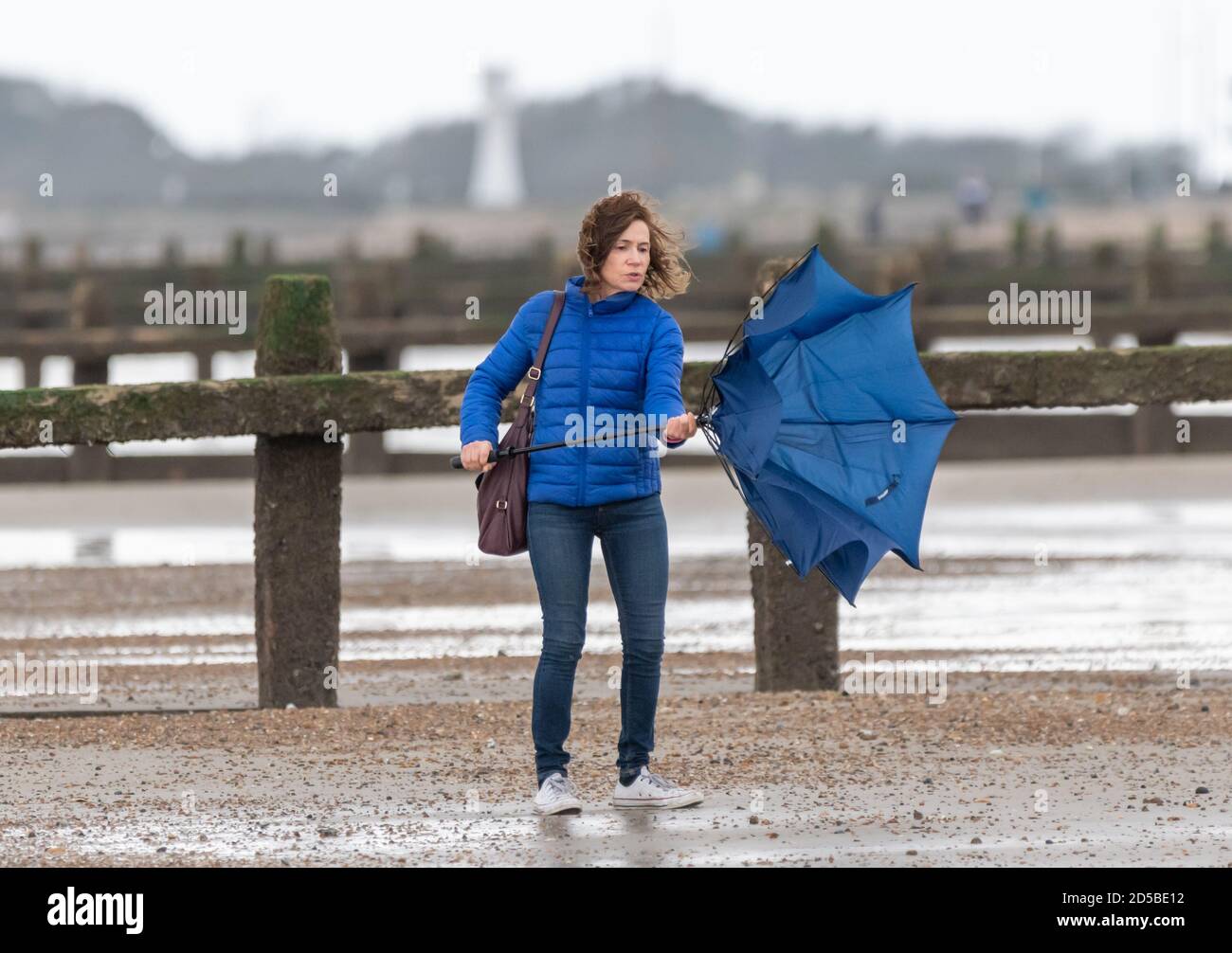 Lady struggling to put up umbrella while walking on a beach in wet and windy weather in the UK. Putting brolly up. Stock Photo