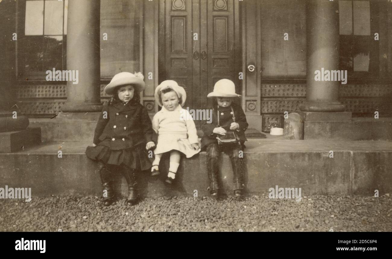 WW! era early 1900's photograph of 3 young children wearing hats sitting on the steps of grand house, circa 1915, one of them is looking at a camera  the children are wearing double breasted coats and puttees protect their shoes, U.K Stock Photo