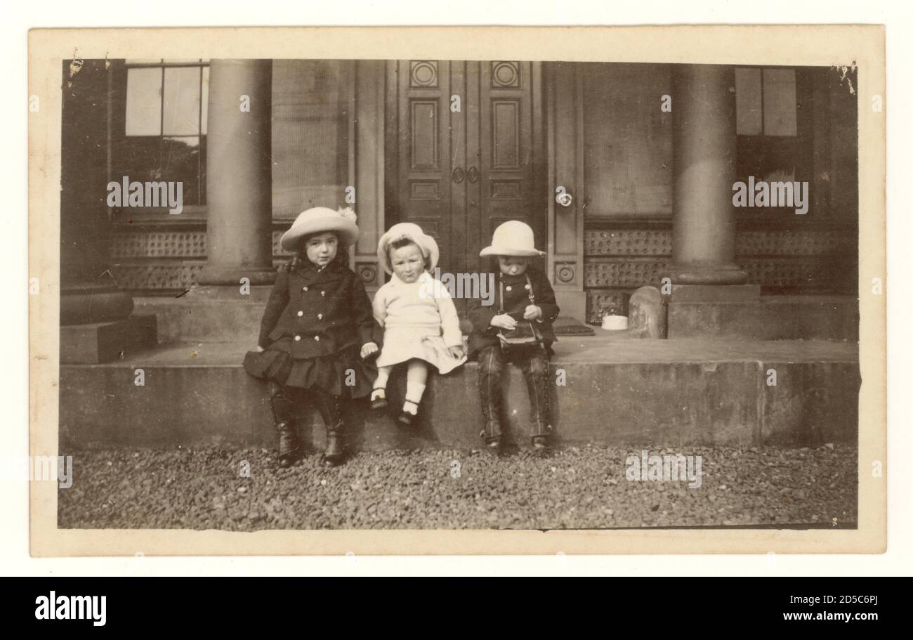 WW1 era early 1900's photograph of 3 young children wearing hats sitting on the steps of grand house, circa 1915, one of them is looking at a camera  the children are wearing double breasted coats and puttees protect their shoes, U.K Stock Photo