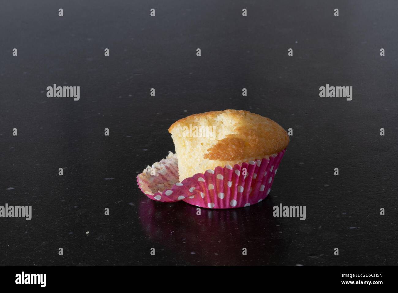 Taking a bite from a plein cupcake, breaking a diet. pink cup with white dots. Dark gray background, kitchen table top Stock Photo