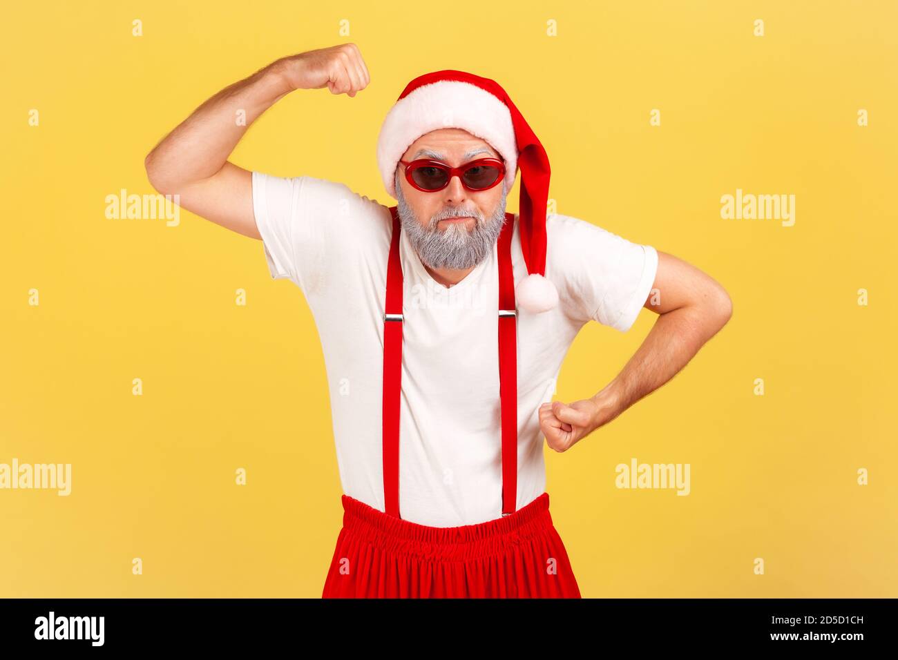 Funny grumpy man in santa claus hat and pants with suspenders showing his arm muscles seriously looking at camera through red stylish sunglasses. Indo Stock Photo