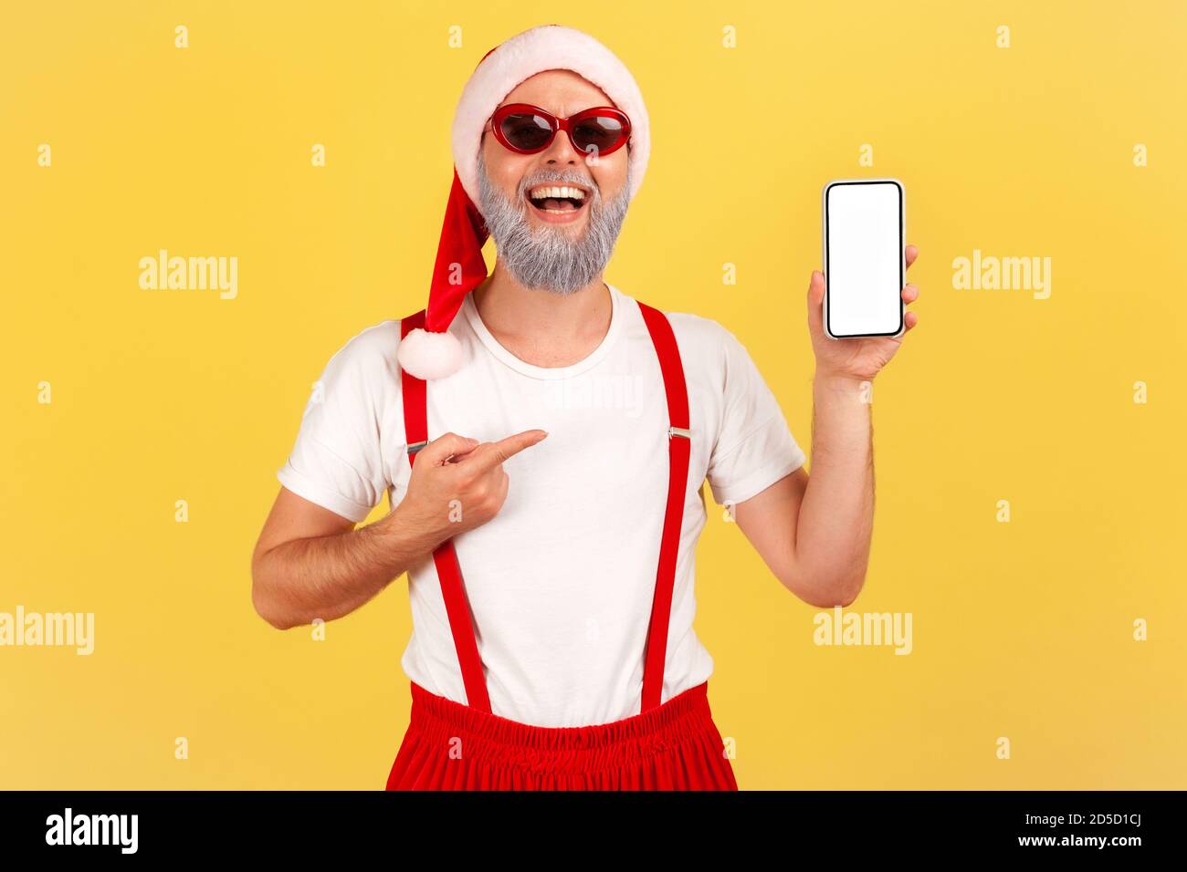 Happy smiling man with gray beard in sunglasses and santa claus hat pointing finger at smartphone with empty white display, online application. Indoor Stock Photo