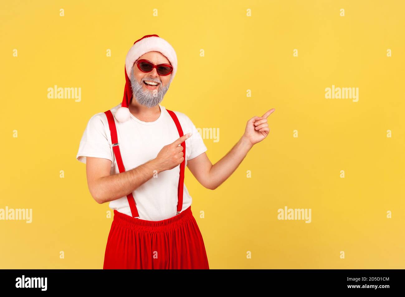 Smiling positive gray bearded man in santa claus costume pointing fingers on empty space, paying attention at advertisement place. Indoor studio shot Stock Photo