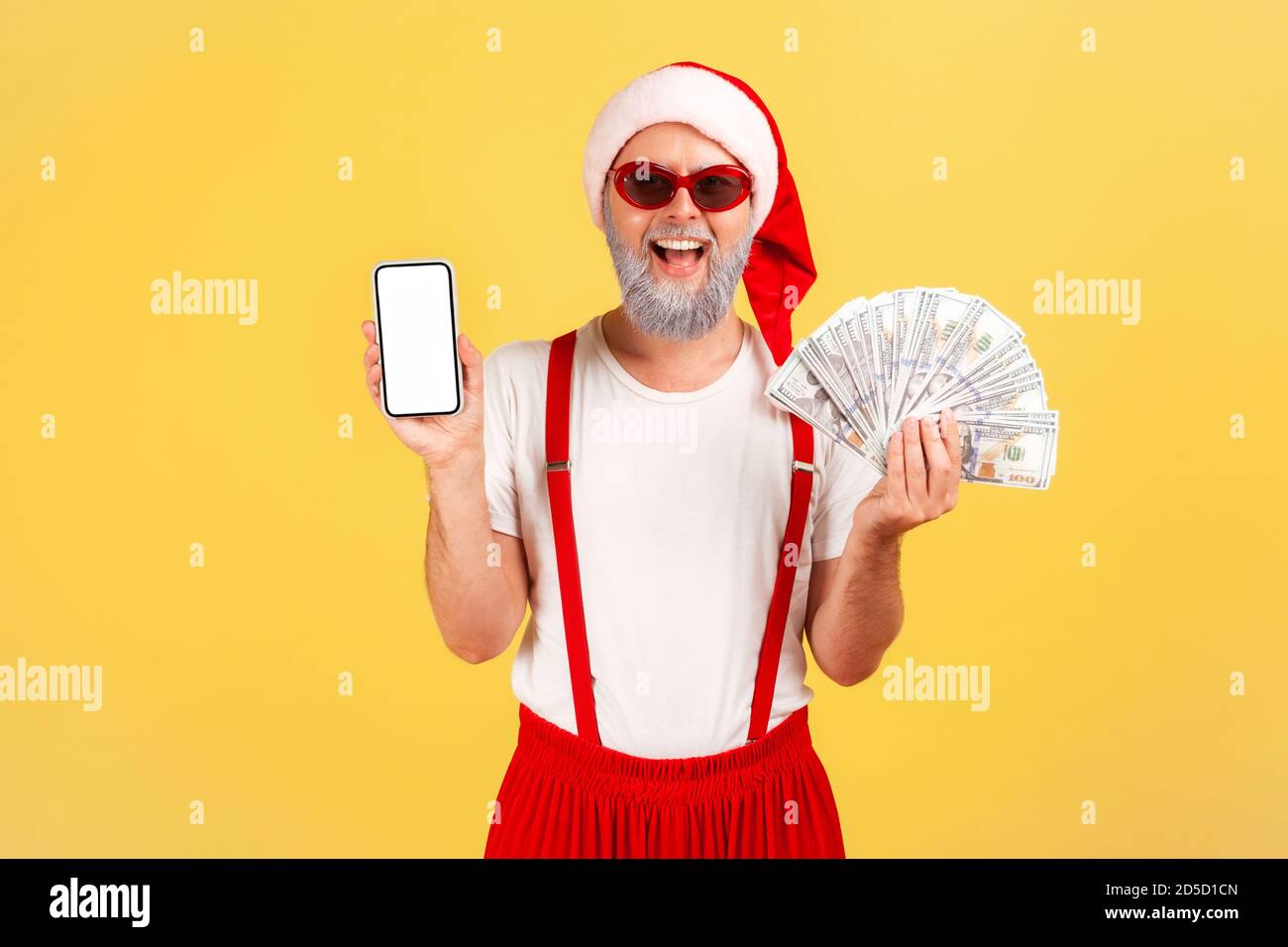 Happy smiling man in santa claus costume and stylish red sunglasses holding fan of dollars and showing smartphone with empty screen, betting app. Indo Stock Photo
