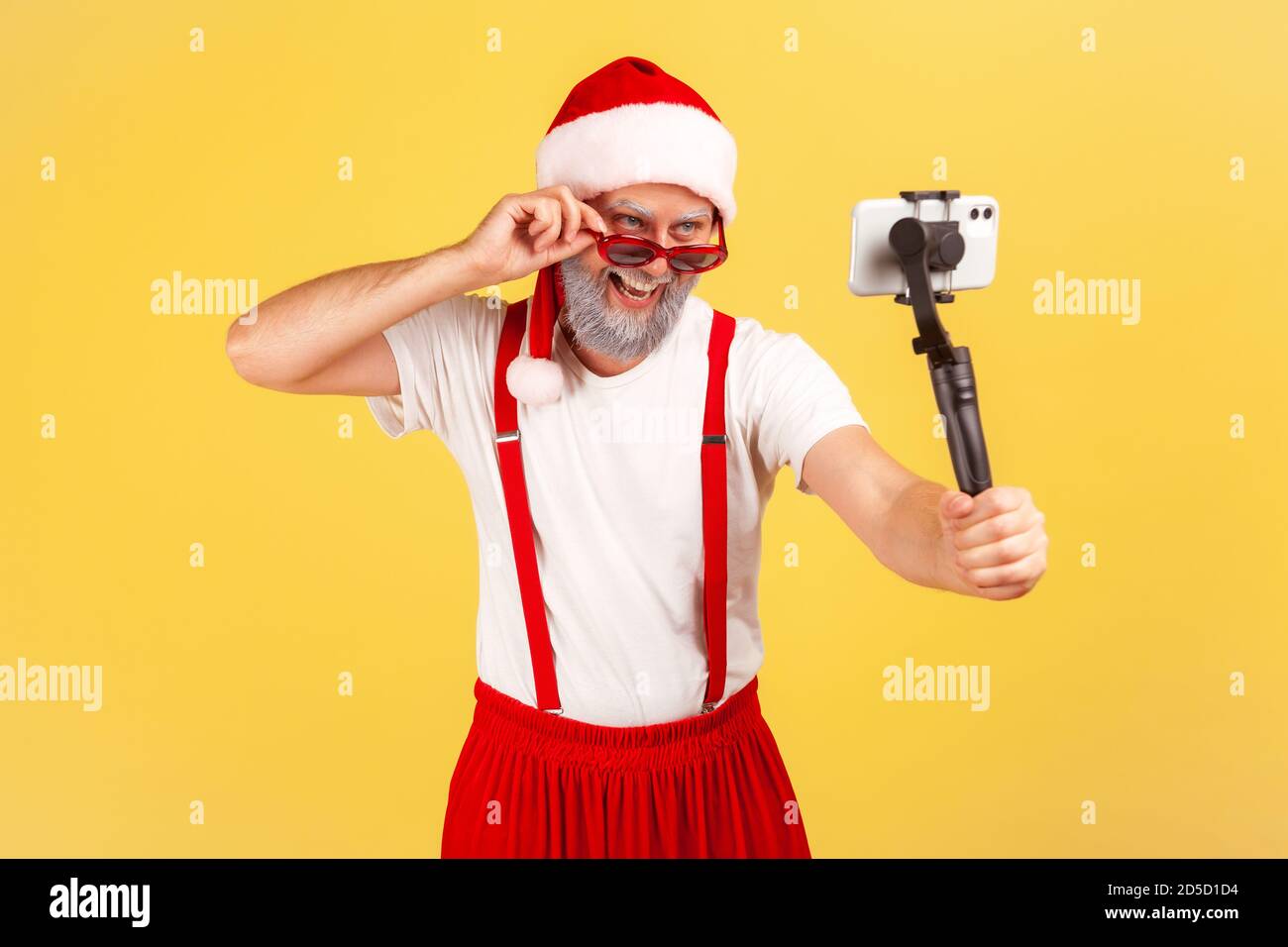 Handsome gray bearded man in santa claus costume holding smartphone on stick posing to make photo or record video, blogging. Indoor studio shot isolat Stock Photo