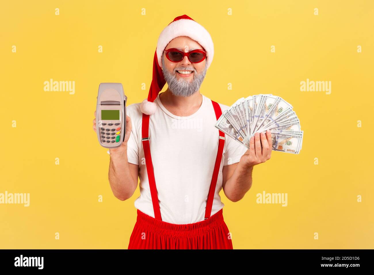 Positive adult man in sunglasses and santa claus hat holding pos terminal and dollar banknotes smiling at camera, choosing cashless payments. Indoor s Stock Photo