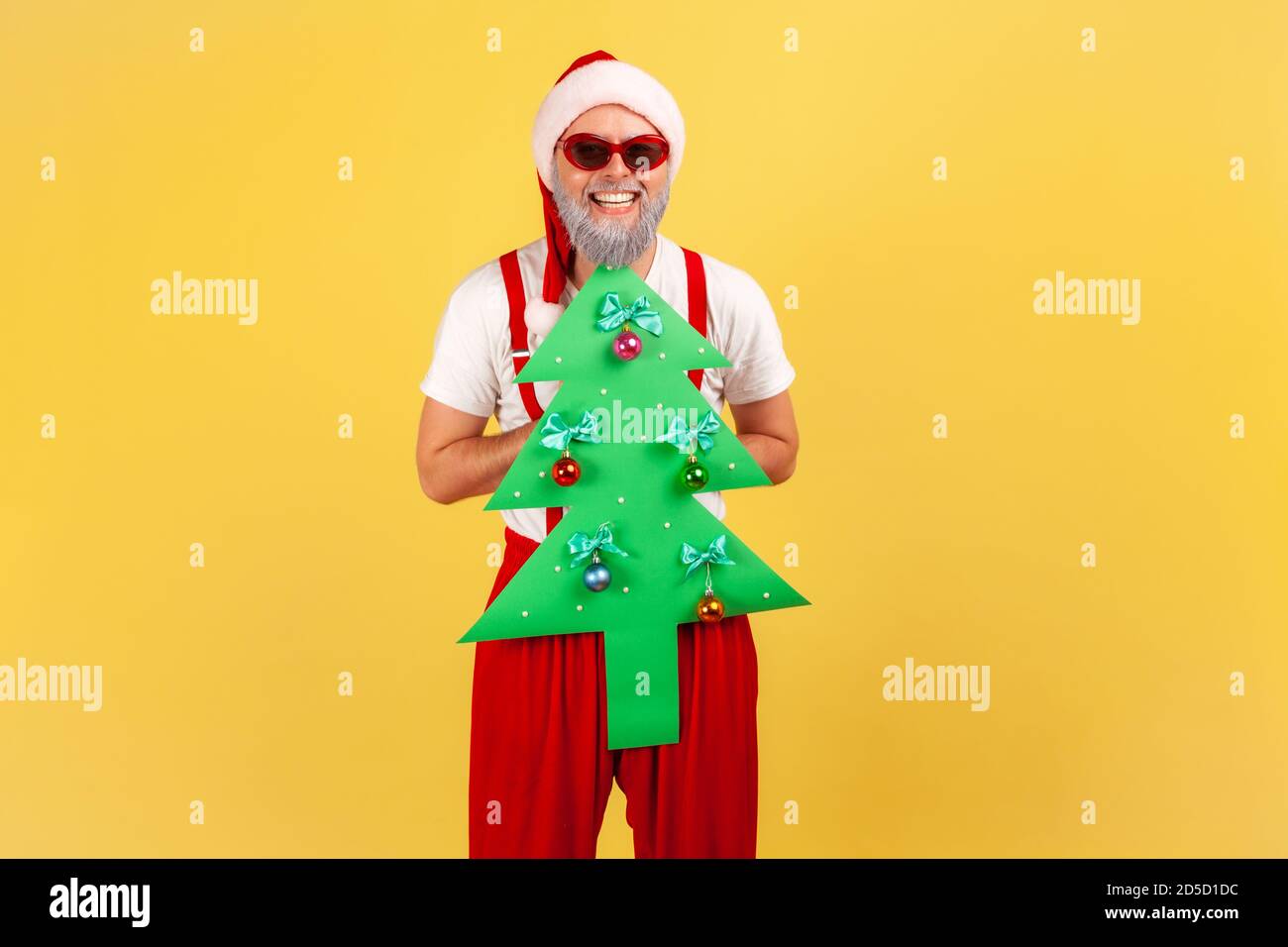 Extremely happy elderly man in santa claus costume and stylish sunglasses holding paper christmas tree looking at camera with toothy smile. Indoor stu Stock Photo