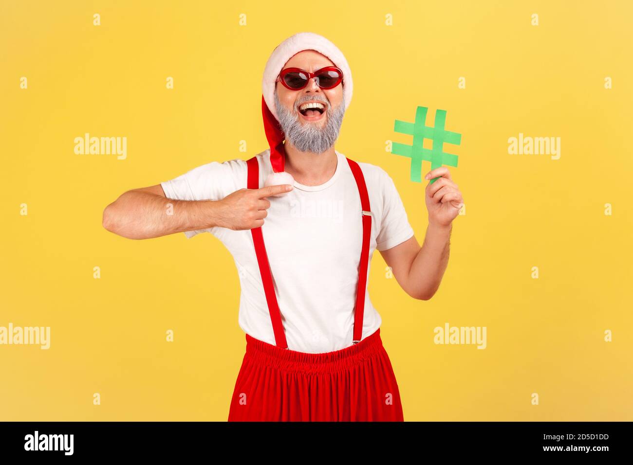 Happy smiling gray bearded man in sunglasses and santa claus hat pointing at green hashtag symbol in his hand, tagging holiday posts. Indoor studio sh Stock Photo