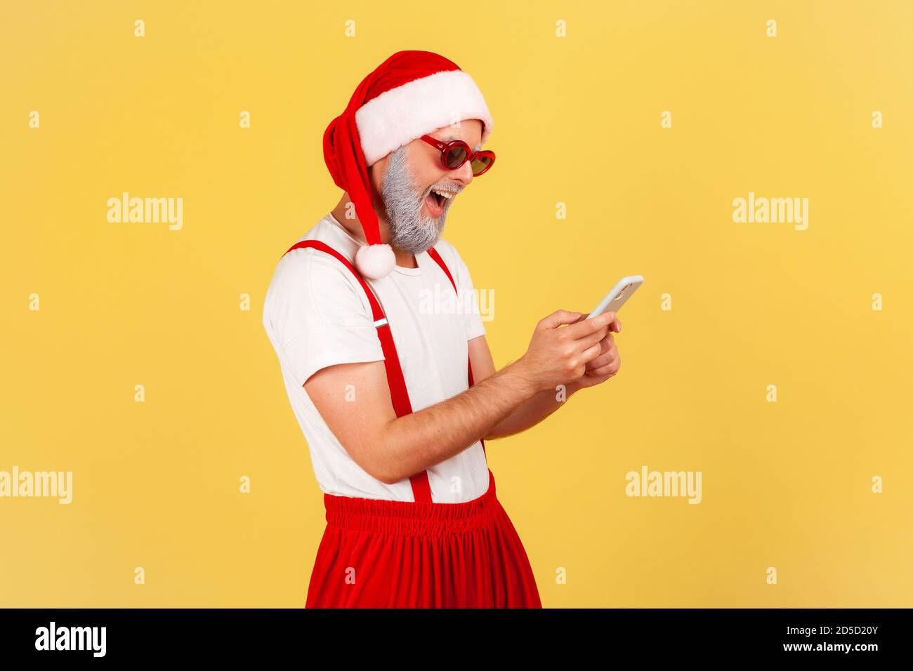Happy funny man in santa claus costume and stylish sunglasses typing on smartphone and smiling, reading pleasant news. Indoor studio shot isolated on Stock Photo