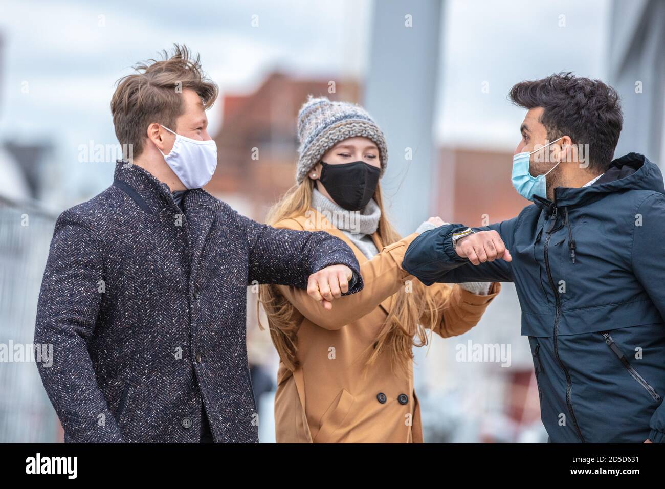 Zwei Männer und eine Frau begrüssen sich mit dem Ellenbogen. Corona-Zeit mit alltäglichen Masken in der kalten Jahreszeit in der Stadt. Stock Photo