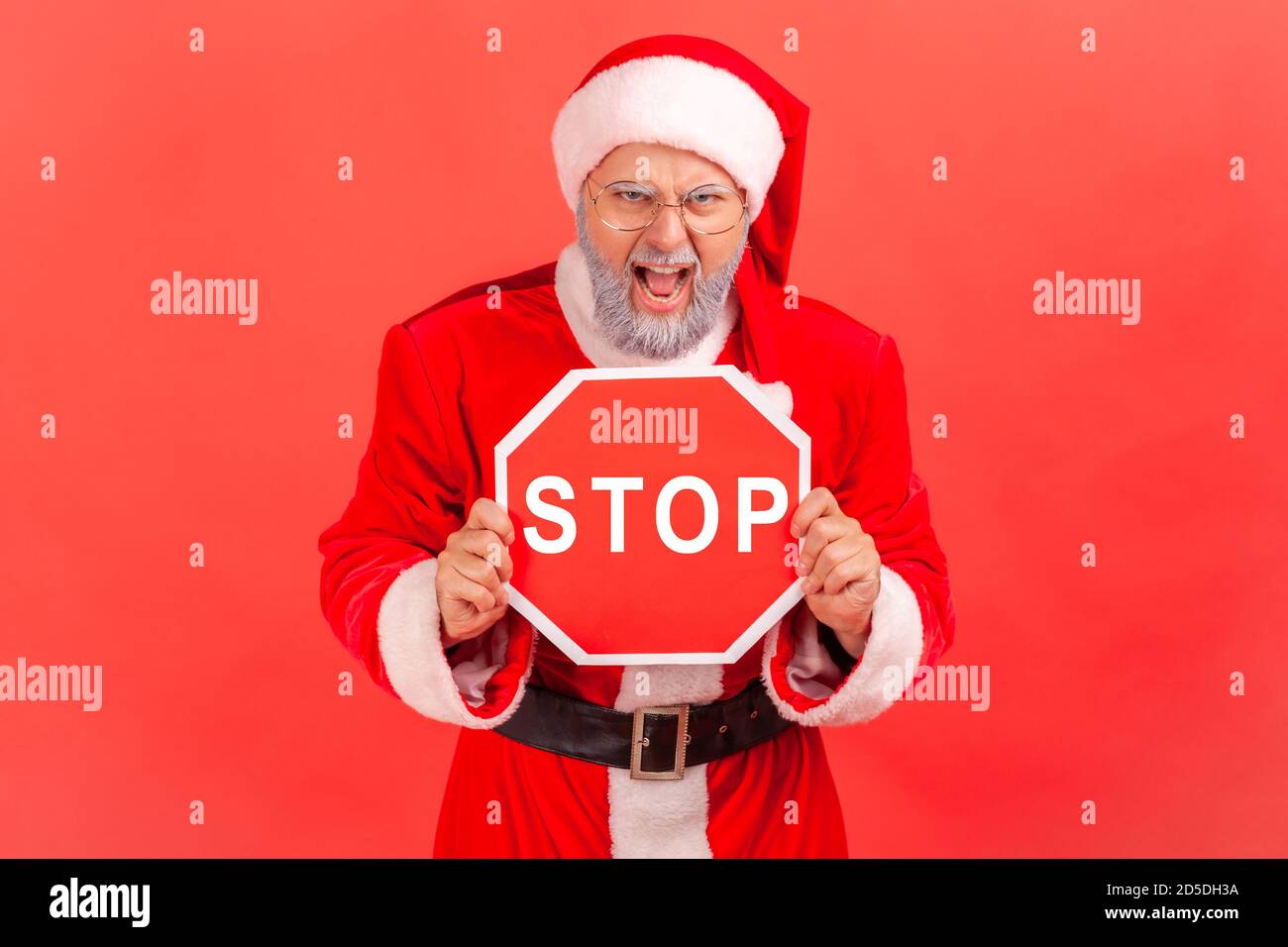 Angry gray bearded man in eyeglasses and santa claus costume shouting holding stop sign, no way, warning about holidays celebration. Indoor studio sho Stock Photo