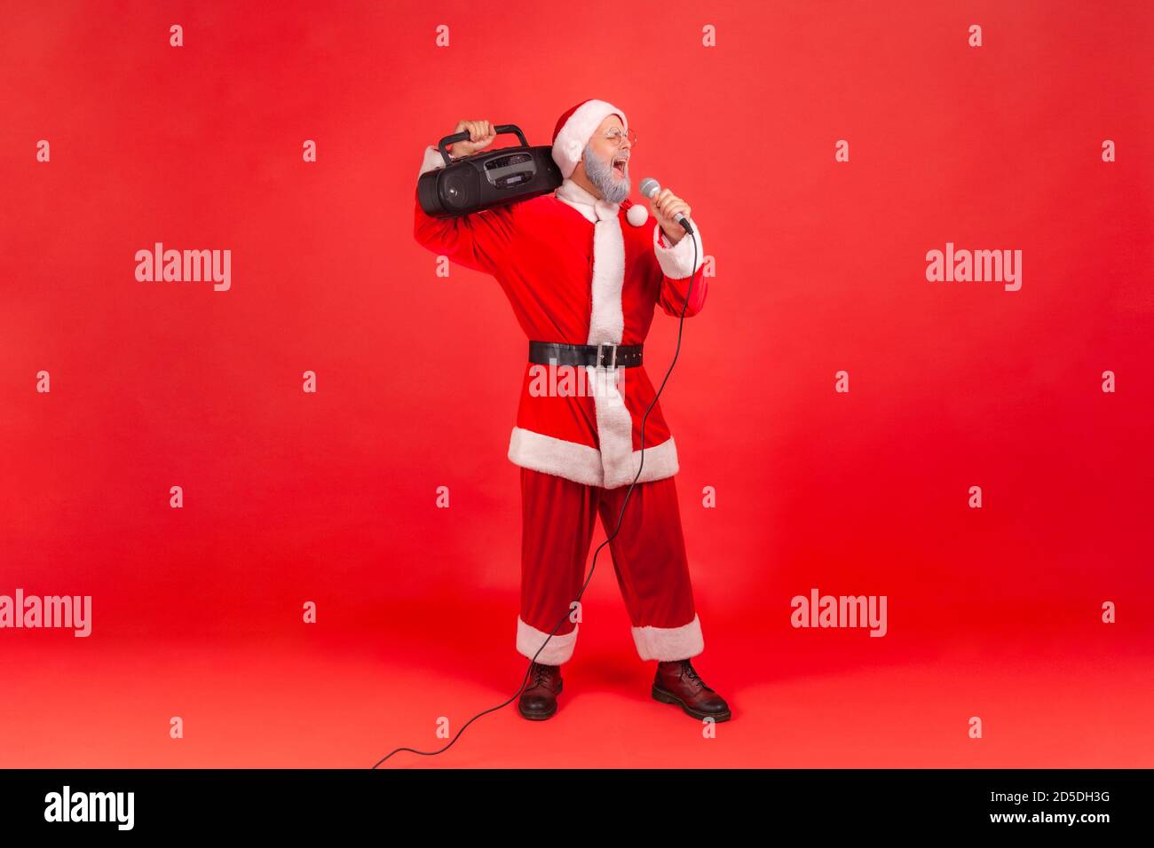 Full length positive cheerful man in santa claus costume singing into microphone holding tape recorder on shoulder, holidays karaoke. Indoor studio sh Stock Photo