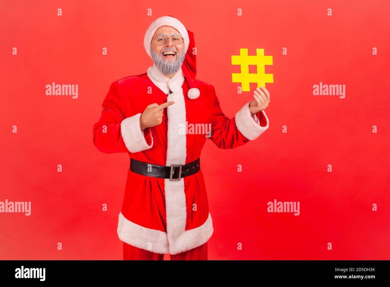 Positive happy gray bearded man in eyeglasses and santa claus costume pointing finger at yellow hashtag symbol in his hand, tagging holiday posts. Ind Stock Photo