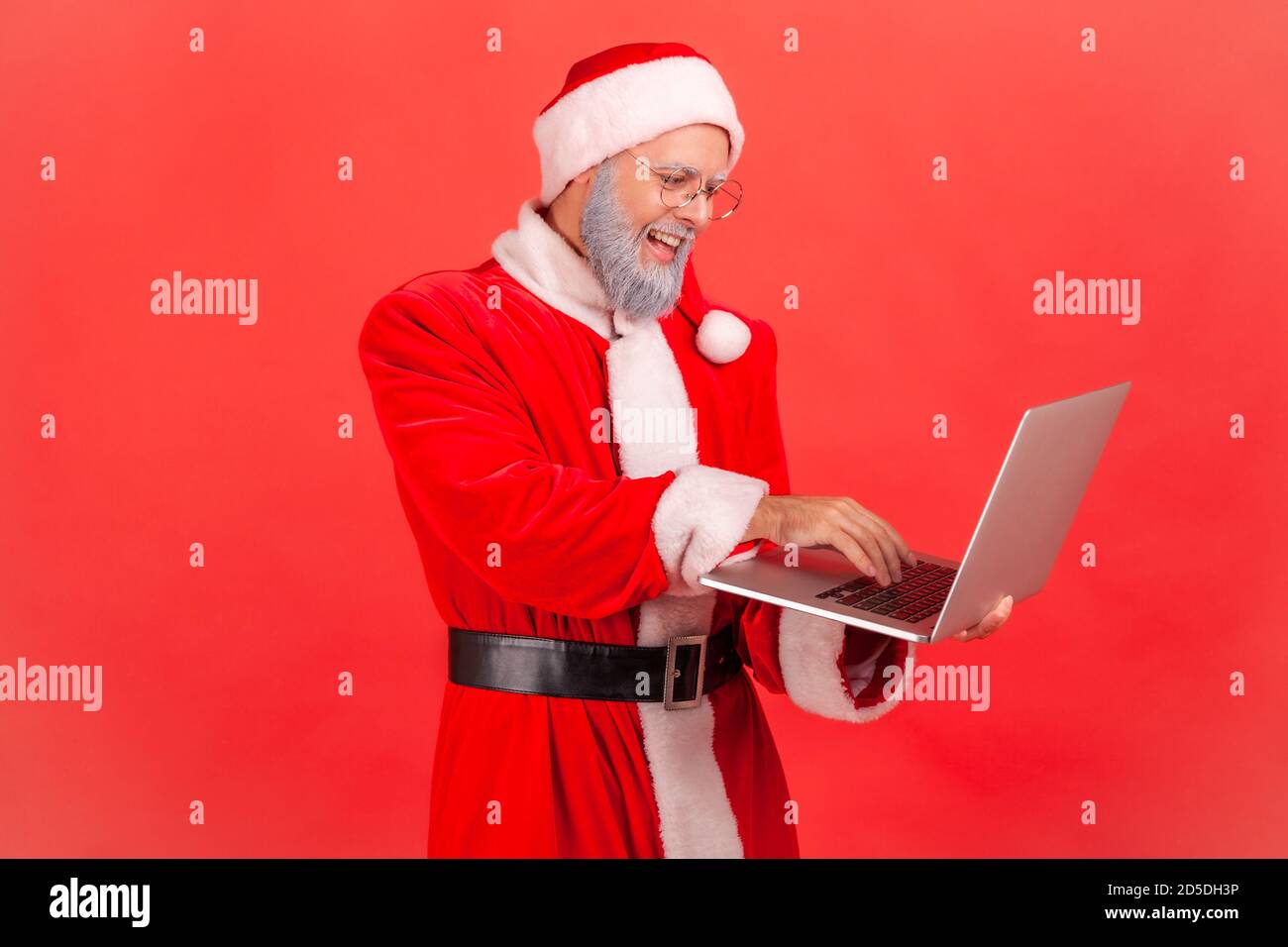 Smiling cheerful man in eyeglasses and santa claus costume working on laptop, sending holidays greeting cards. Indoor studio shot isolated on red back Stock Photo