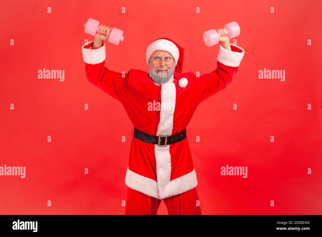 Positive funny elderly man in santa claus costume and eyeglasses raising dumbbells and fooling face, preparing for winter holidays. Indoor studio shot Stock Photo