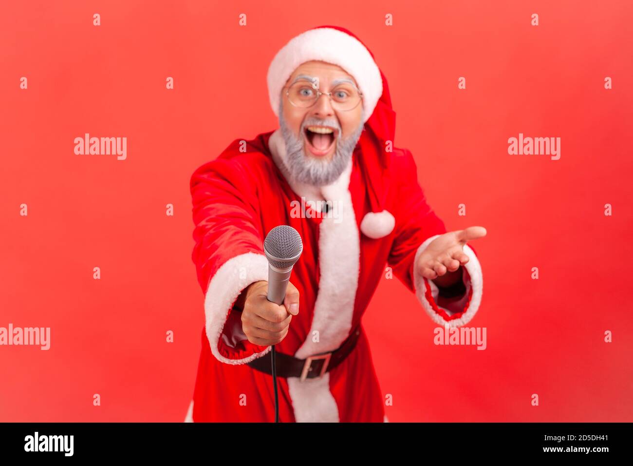 Excited positive gray bearded man in santa claus costume holding microphone asking you to sing or say congratulation. Indoor studio shot isolated on r Stock Photo