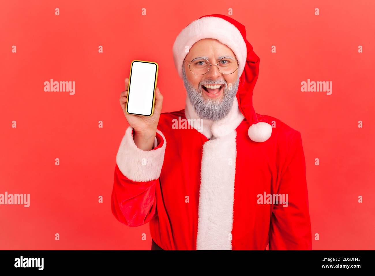 Happy gray bearded santa claus holding and showing smartphone with white display looking at camera with toothy smile. Indoor studio shot isolated on r Stock Photo
