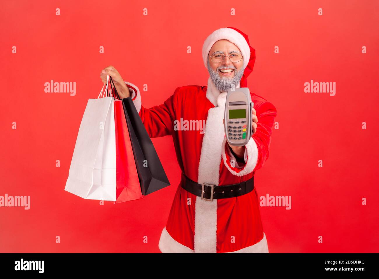 Positive gray bearded man in eyeglasses and santa claus costume holding and showing paper shopping bags and payment terminal, contactless payments. In Stock Photo