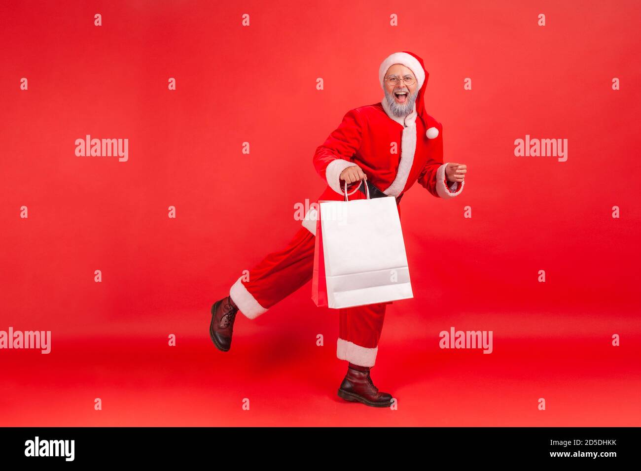 Full length happy satisfied santa claus holding paper bags, doing holidays shopping, enjoying christmas sale. Indoor studio shot isolated on red backg Stock Photo