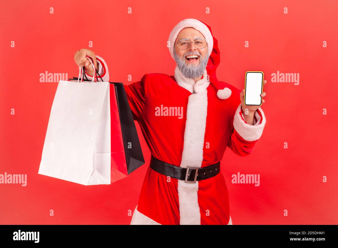 Smiling positive gray bearded santa claus holding and showing paper bags and empty screen smartphone, shopping online. Indoor studio shot isolated on Stock Photo