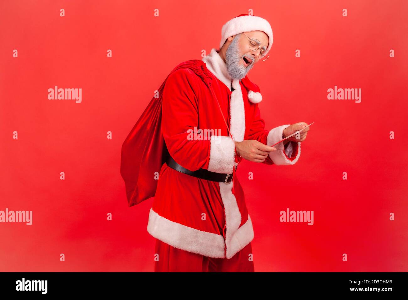 Kind gray bearded santa claus with big red bag reading letters with presents requests and greeting cards, magic of winter holidays. Indoor studio shot Stock Photo