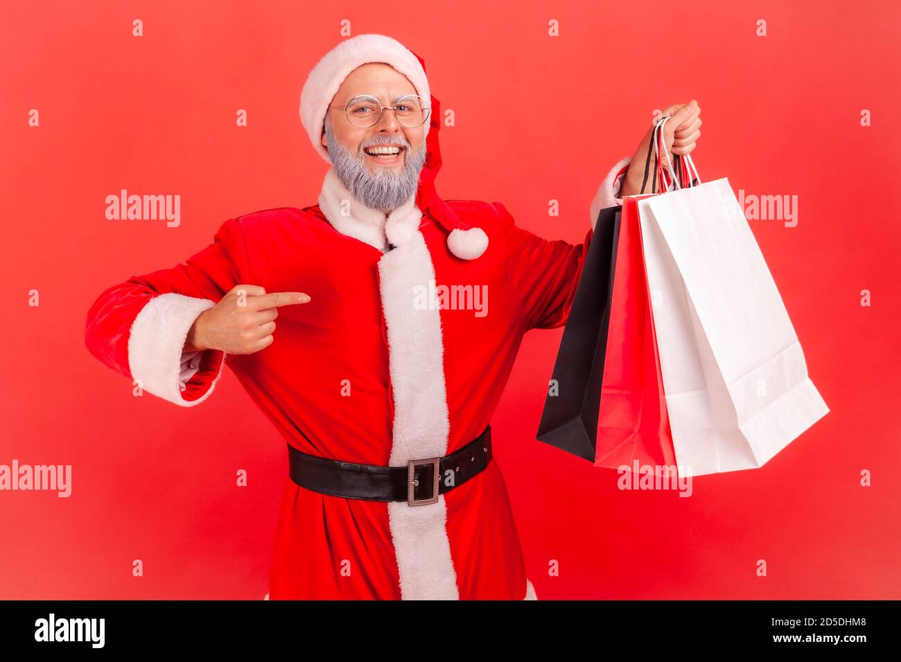 Smiling elderly gray bearded man in santa claus costume pointing finger at paper shopping bags in his hand, doing holidays purchase. Indoor studio sho Stock Photo
