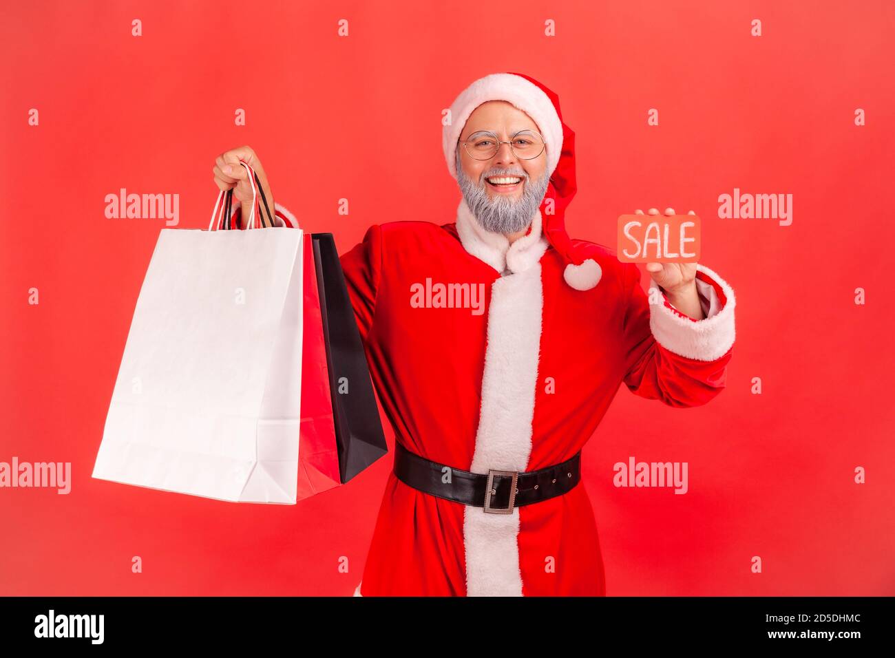 Happy joyful santa claus holding and showing sale card and colorful paper bags looking at camera with toothy smile, satisfied with holidays discounts. Stock Photo