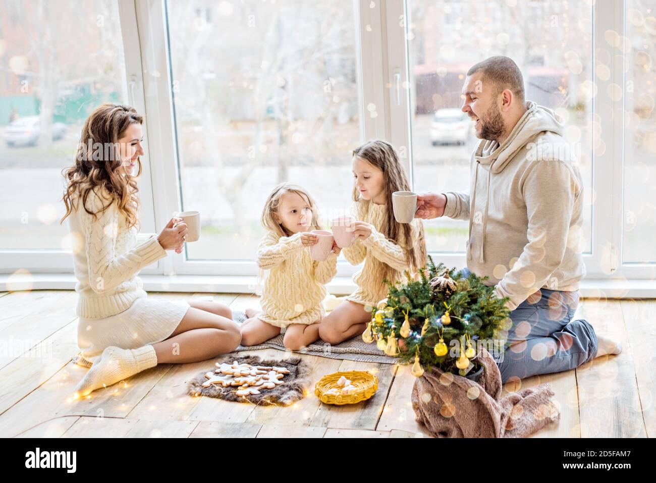 Happy family in white sweaters drinks cocoa by the panoramic window. Parents and daughters are holding cups in their hands, on a plate are gingerbread Stock Photo