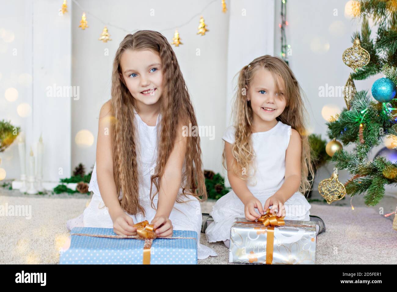 Two little blonde girls in white dresses are playing by the fireplace ...