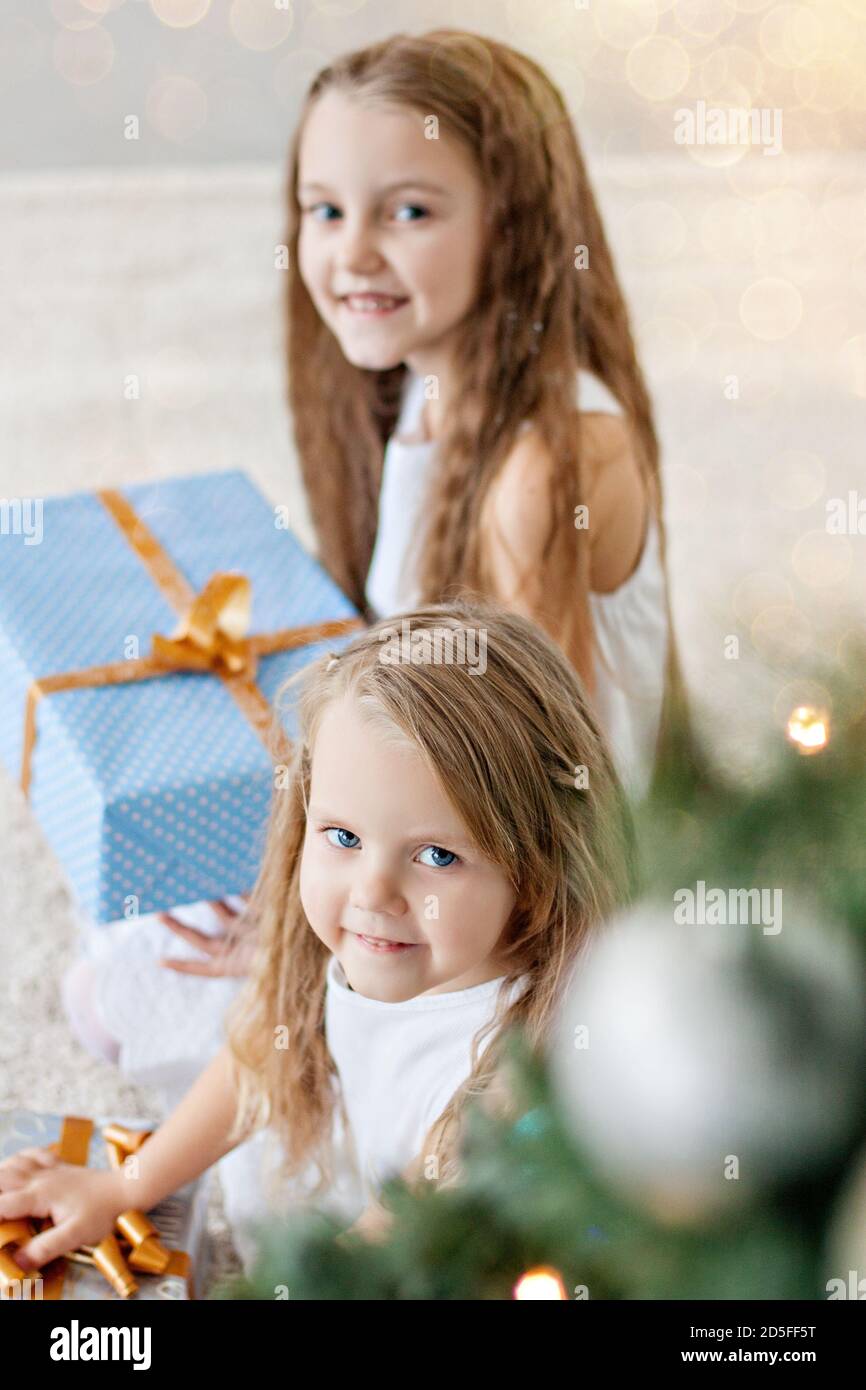 Two little blonde girls in white dresses are playing by the fireplace and Christmas tree with boxes. Sisters have fun, hug, laugh, unfold gifts Stock Photo