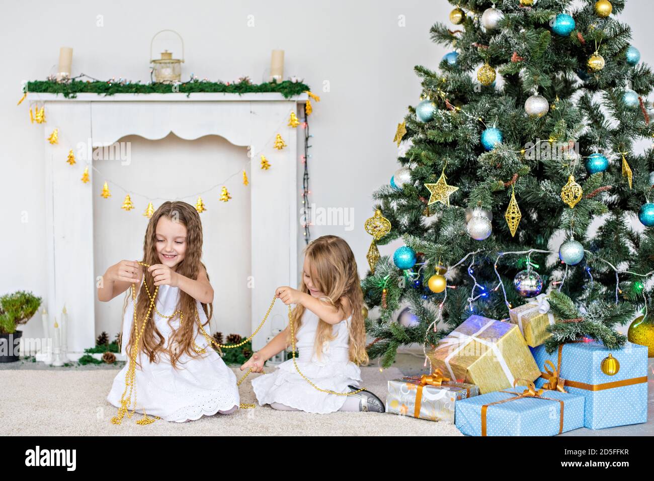 Two little blonde girls in white dresses are playing by the fireplace and Christmas tree with boxes. Sisters have fun, hug, laugh, unfold gifts Stock Photo