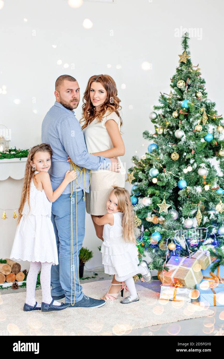 Happy family, parents hugging with their daughters by the white fireplace with an elegant Christmas tree in garlands of lights. New year copy space Stock Photo