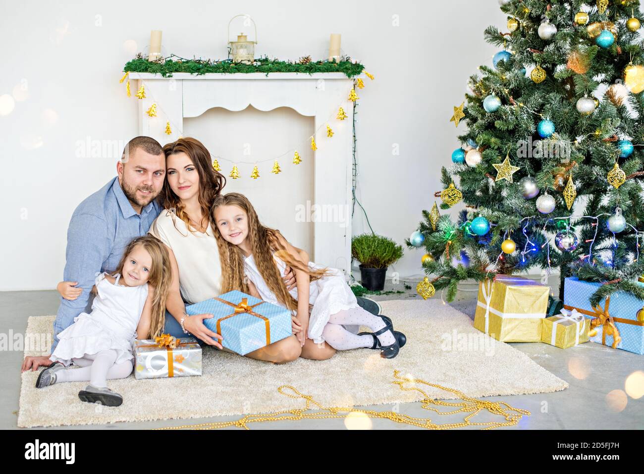 Happy family are holding boxes with gifts in front of a white fireplace with an elegant Christmas tree in garlands of lights. Young father and beautif Stock Photo