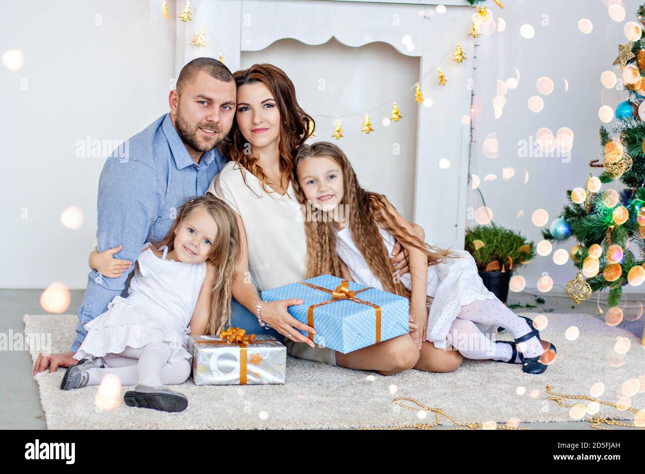Happy family are holding boxes with gifts in front of a white fireplace with an elegant Christmas tree in garlands of lights. Young father and beautif Stock Photo