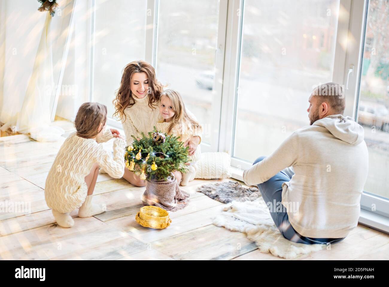 A beautiful young couple family and their two daughters are having fun celebrating New Year. Christmas tree in a pot, large panoramic window and festi Stock Photo