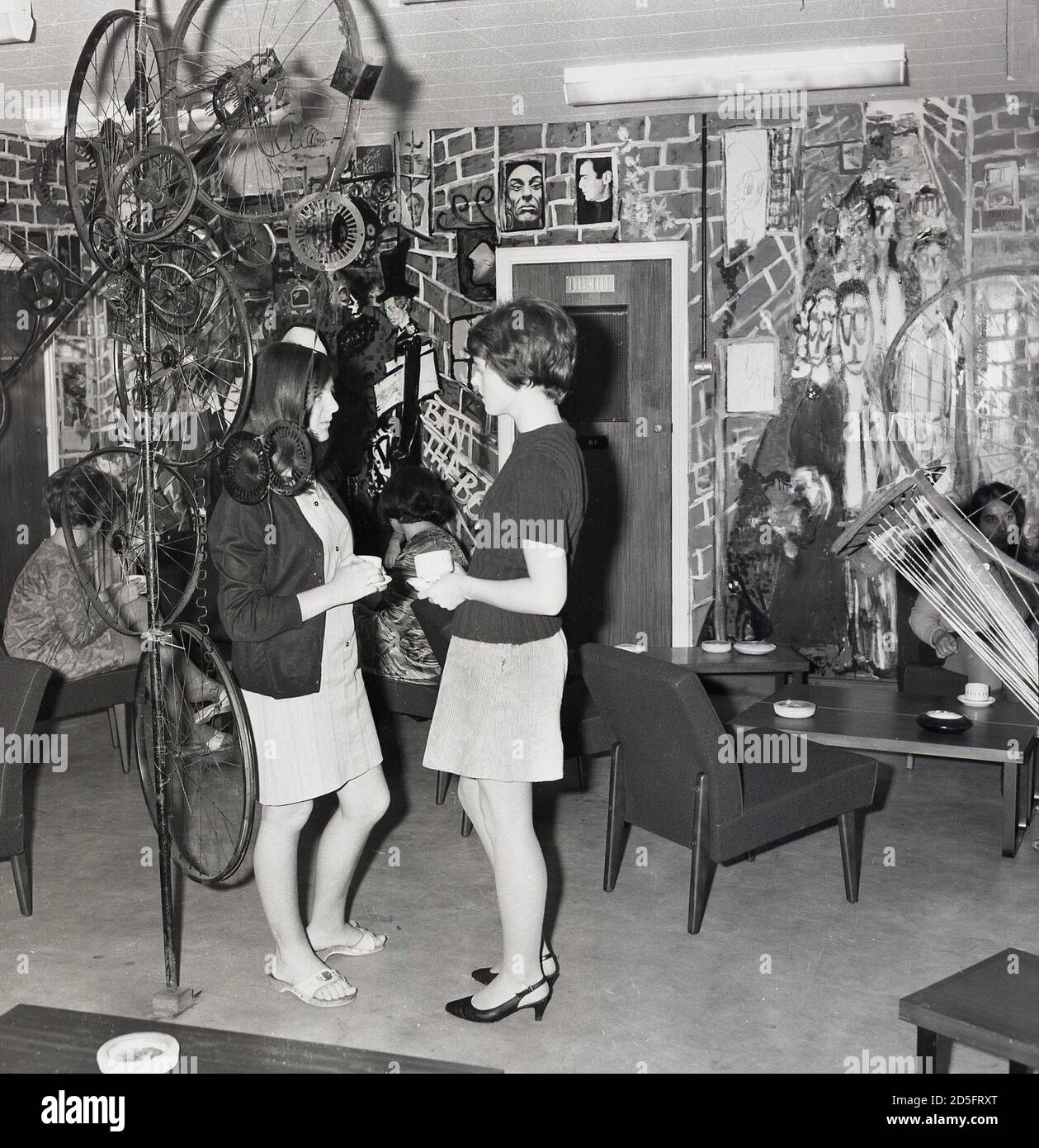 1970s, historical, two young women standing in a student coffee lounge beside an artwork made from old bicycle wheels, Southeast London, England, UK. Stock Photo