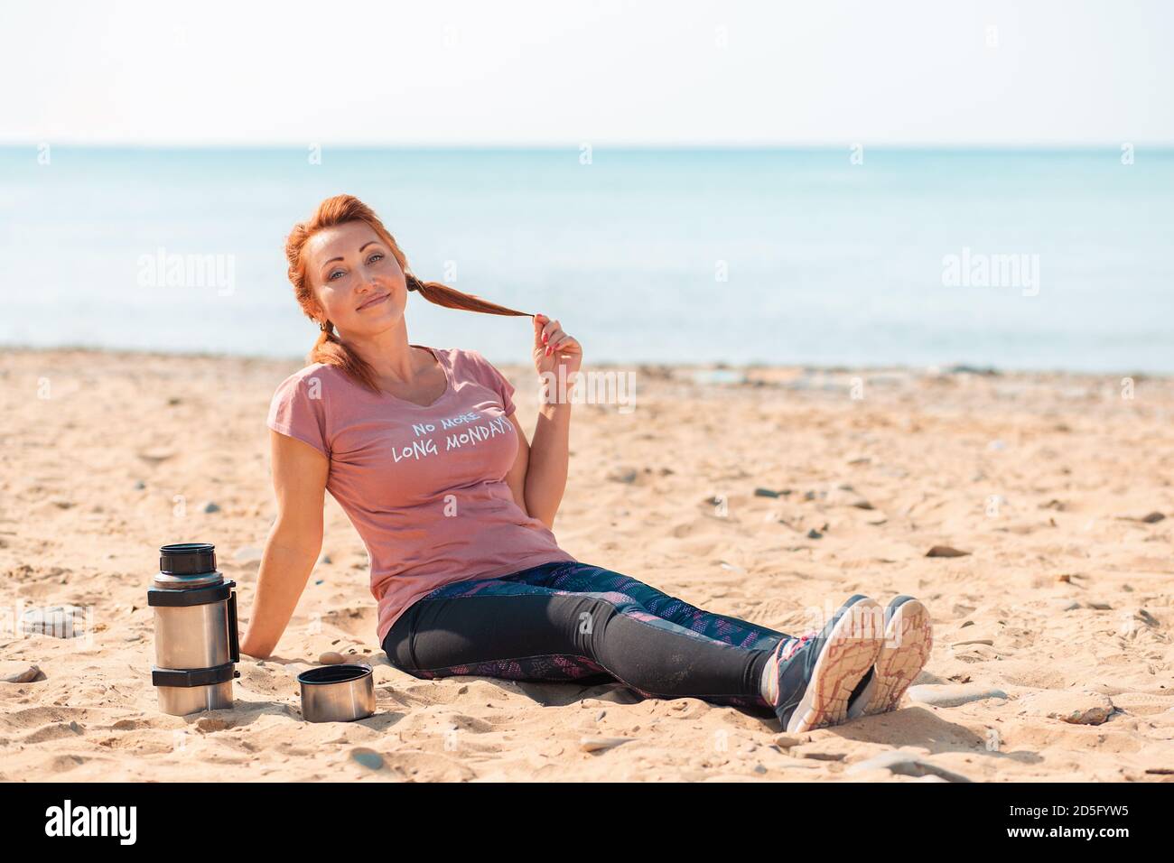 Smiling adult woman in sports clothes, sitting on the sand. Next to it is a bottle of sports nutrition. Sea in the background. The concept of sports l Stock Photo