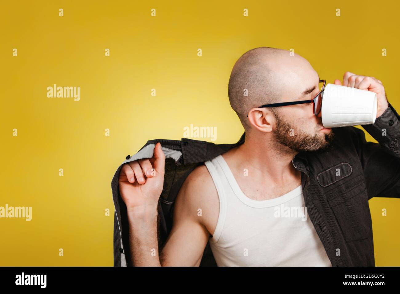 The concept of morning awakening and productivity. A bald, bearded man wearing glasses, wearing a shirt and drinking coffee or tea from a mug at the s Stock Photo