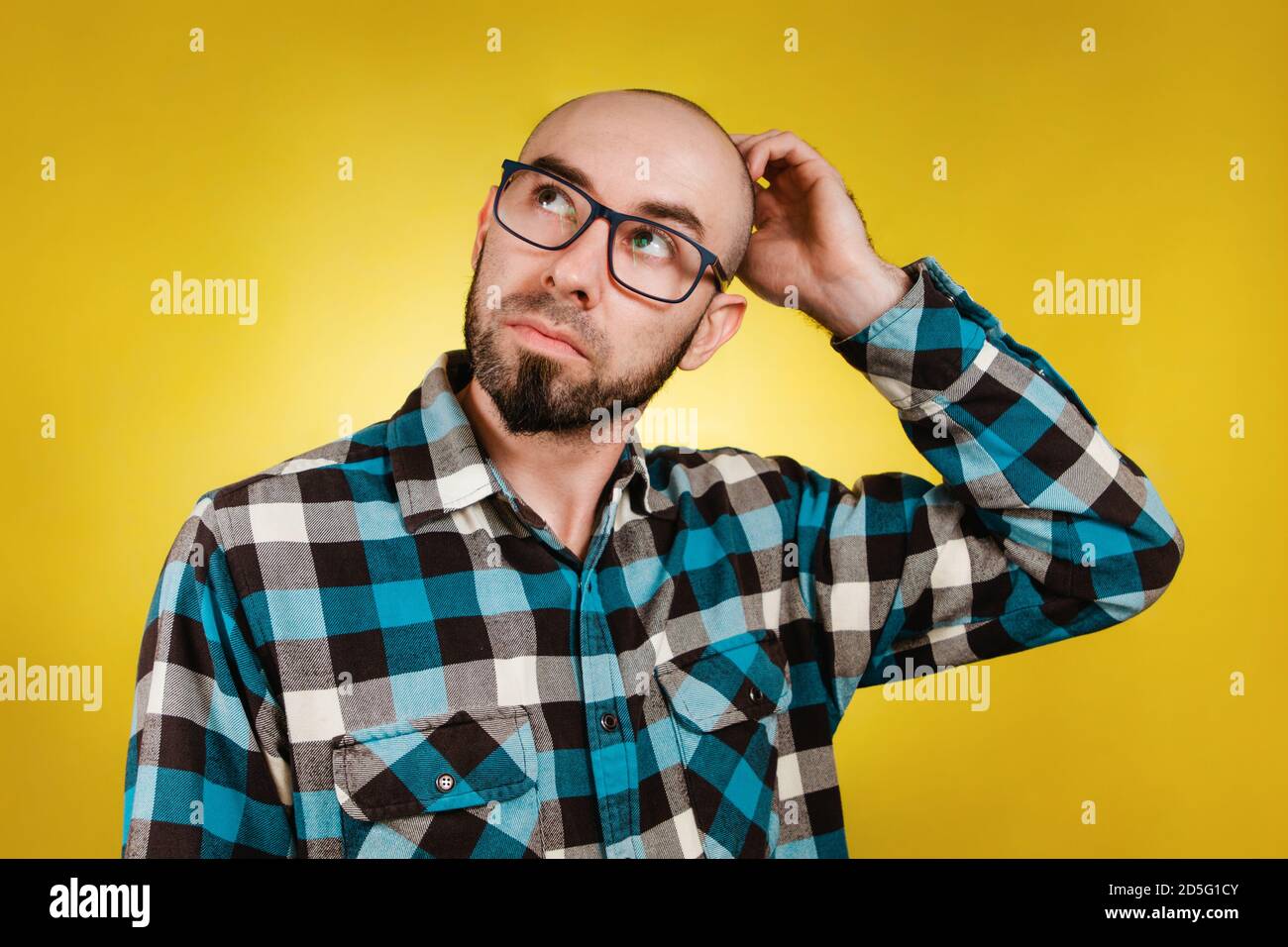 People and emotions. A bald, bearded man in glasses and a blue plaid shirt, scratching his head for a solution or an idea. Yellow background. Copy spa Stock Photo