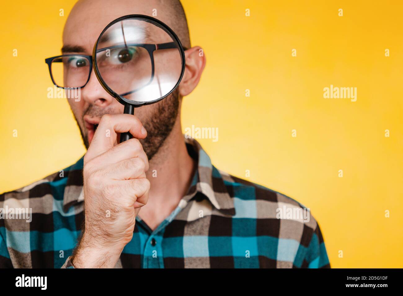 The concept of finding and solving problems. A bald man with a beard and glasses, wearing a blue checked shirt, looks through a magnifying glass. Eye Stock Photo