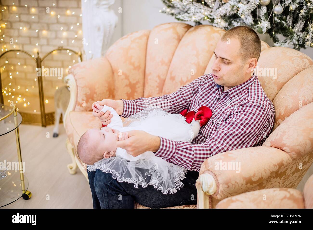 A young man holds a baby in his arms. Father plays with a girl in a white dress, hugs her, kisses against the background of festive Christmas trees Stock Photo