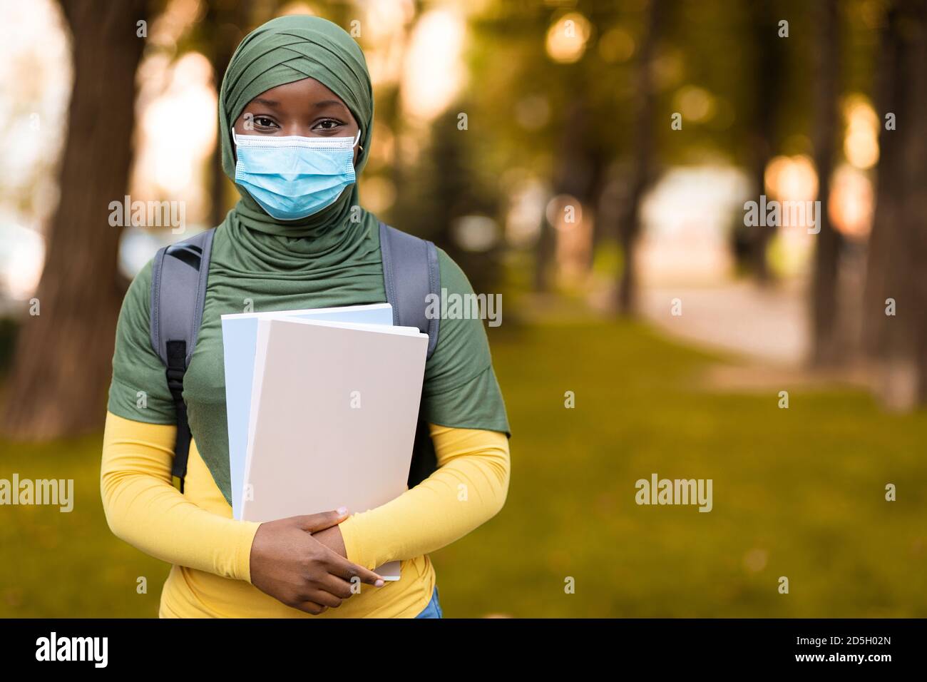 Black muslim female student wearing medical mask outdoors, holding backpack and books Stock Photo