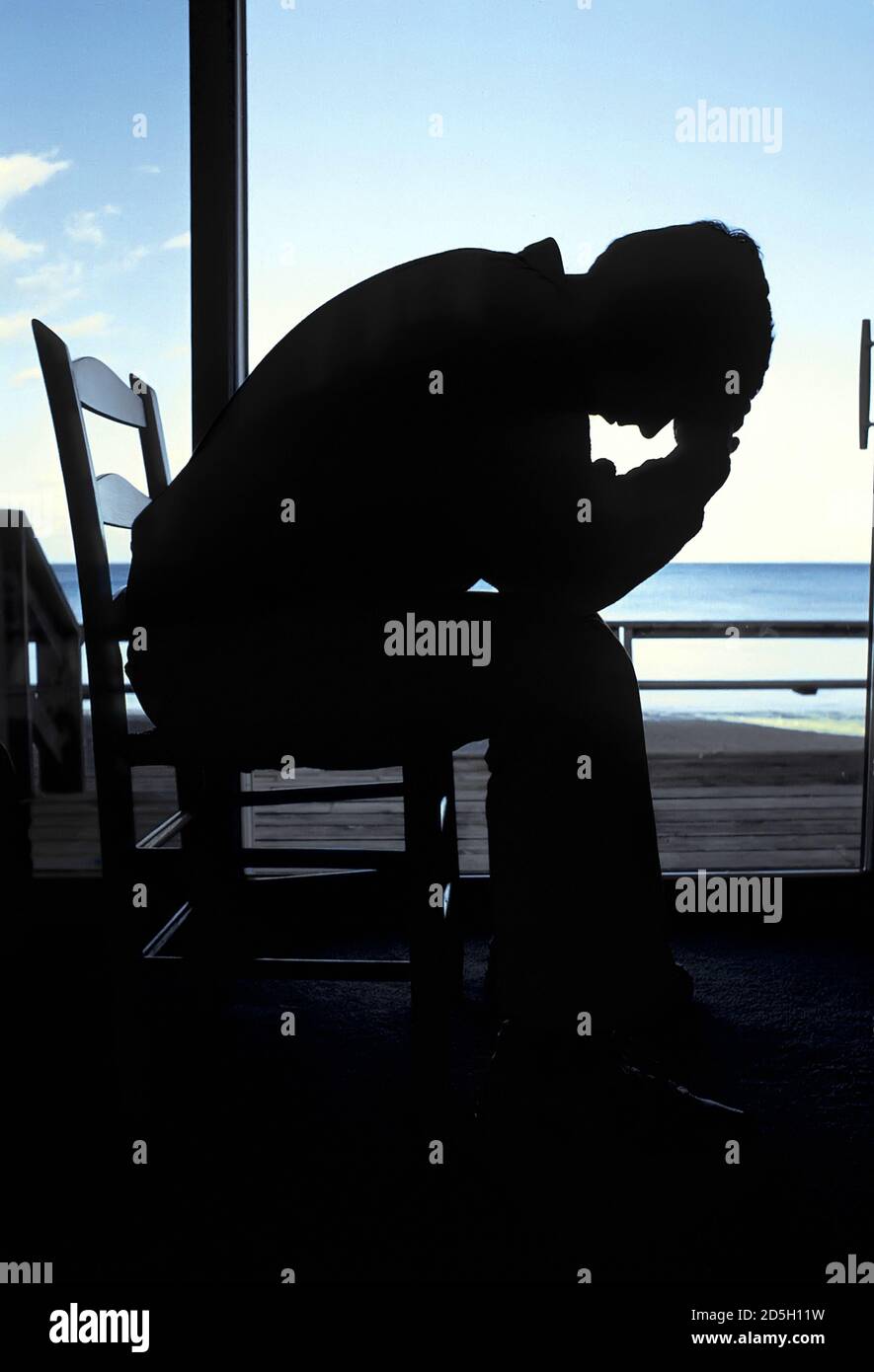 Silhouette of an adult male sitting in a chair with head in hand in deep depression comtemplating suicide Stock Photo