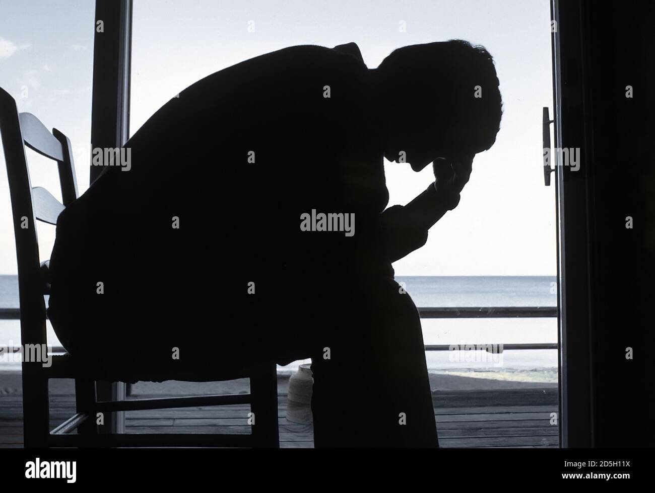 Silhouette of an adult male sitting in a chair with head in hand in deep depression comtemplating suicide Stock Photo
