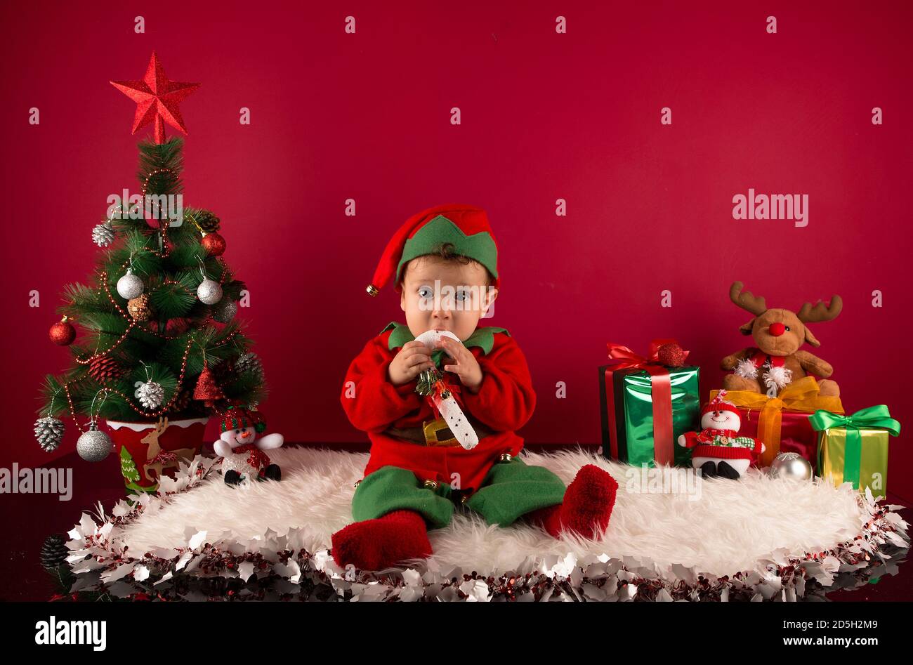 Sweet one-year-old boy dressed as an elf, in Christmas decoration, with gifts and typical tree Stock Photo